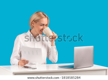 Happy female secretary or office employee with documents, coffee and laptop at workplace over blue studio background, copy space. Positive business lady with papers and hot beverage at desk