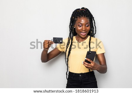 Picture of smiling young african lady standing isolated over white background. Looking aside while using phone and holding debit card.