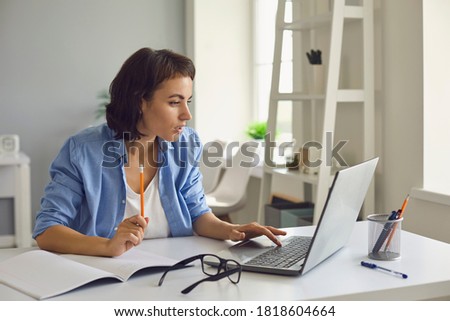 Young woman talking to somebody and making notes during online conversation, lesson or teleconference on laptop at home. Online communication and elearning concept Royalty-Free Stock Photo #1818604664