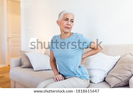 Unhappy grey haired mature woman with closed eyes touching, massaging back, tired sad middle aged grandmother suffering from backache, pinched nerve, sitting in living room at home alone
