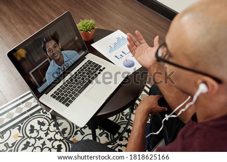 Young businessman sitting at small table with financial report and video calling his Vietnamese colleague Royalty-Free Stock Photo #1818625166