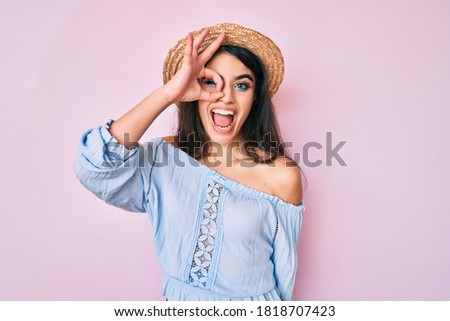 Brunette teenager girl wearing summer hat smiling happy doing ok sign with hand on eye looking through fingers 