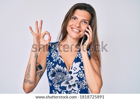 Beautiful caucasian woman having conversation talking on the smartphone doing ok sign with fingers, smiling friendly gesturing excellent symbol 