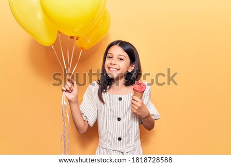 Adorable hispanic child girl smiling happy. Standing with smile on face holding balloons eating ice cream over isolated yellow background