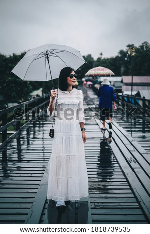 Asia woman traveler in the white dress holding white umbrella standing on the wood bridge when the rain are fall.