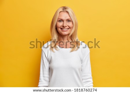 Optimistic glad blonde middle aged woman smiles toothily happy to spend free time in family circle wears casual white jumper isolated on yellow background. Happy senior wrinkled lady indoor.