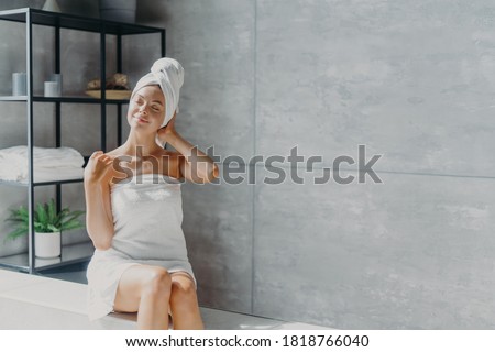 Relaxed young Caucasian female model wears towel wrapped on head, feels refreshed after taking shower, has healthy clean soft skin, poses in cozy bathroom. Women, beauty and hygiene concept. Royalty-Free Stock Photo #1818766040