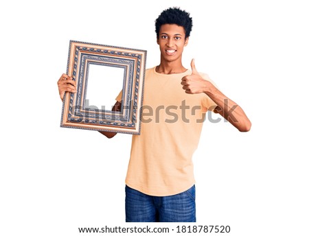 Young african american man holding empty frame smiling happy and positive, thumb up doing excellent and approval sign 
