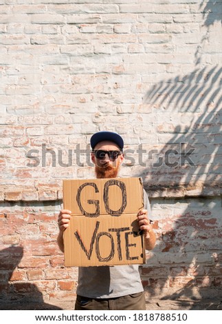 Man shows cardboard with Go Vote sign on brick wall urban background. Voting concept. Make the political choice, use your voice. Bearded guy in sunglasses invite to go to the presidential elections.