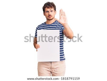 Young handsome man with curly hair holding blank empty banner with open hand doing stop sign with serious and confident expression, defense gesture 