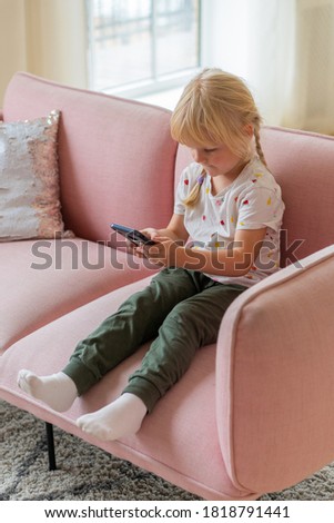 Beautiful little girl playing game or watching video on smartphone mobile. Girl watching cartoons or browsing internet, copy space. Side view portrait of little girl using smartphone while sitting on 