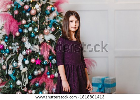 Seven-year-old dark-haired girl in purple dress stands near the elegant Christmas tree indoors