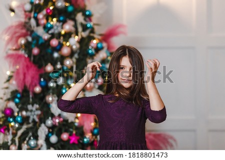 funny Seven-year-old dark-haired girl in purple dress stands near the elegant Christmas tree indoors and looking to camera. straightens her hair