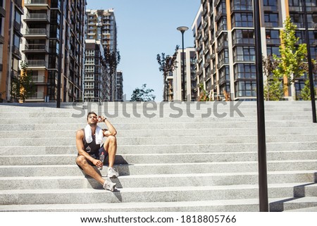 Young man exercising outside. Picture of tired guy after harworking training or exercising. Sit alone on steps and drinking water. White towel around neck. Rest and relax.