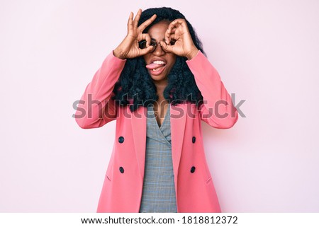 Beautiful african woman wearing business jacket and glasses doing ok gesture like binoculars sticking tongue out, eyes looking through fingers. crazy expression. 