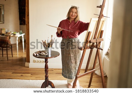 Full length image of stylish barefoot mature female artist in apron painting indoors, standing next to easel and table with many paintbrushes, finishing picture. Art and creative occupation