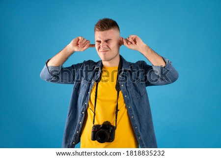 Irritated male traveler with photo camera covering his ears with fingers on blue studio background. Male passenger traveling by plane, suffering from noise during his flight