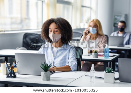 Social distancing at coronavirus outbreak situation. Pretty african american lady in protective mask typing on laptop, working for company support center in office interior with other employees Royalty-Free Stock Photo #1818835325