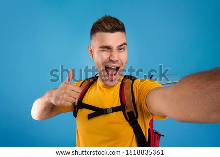 Cool young hipster with tourist equipment taking selfie, winking and showing thumb up gesture over blue studio background. Positive hiker taking funny photo of himself before camping trip