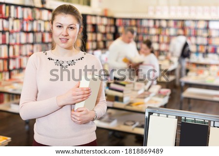 Portrait of attractive young woman holding thick book in bookstore interior