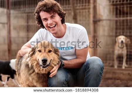 Happy young male volunteer sitting with adorable hairy homeless shepherd dog against cages with animals in shelter Royalty-Free Stock Photo #1818894308