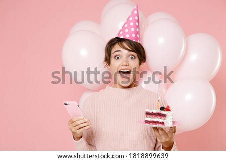 Excited young brunette woman in casual sweater birthday hat celebrating birthday holiday party hold cake with candle using mobile cell phone isolated on pastel pink background studio with air balloons
