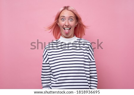 Beautiful young female has pink hair and pleasant appearance, shows tongue, has fun, teasing her friend