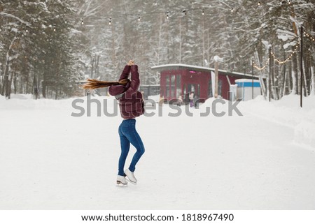 Young girl is skating at an ice rink in a winter park outdoors. Warm clothes, knitted hats, snowdrifts, trees, blurred background.