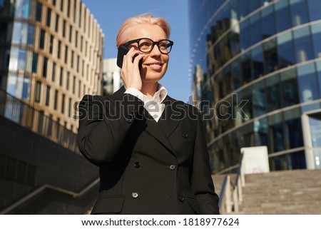 Mature businesswoman in black suit talking on mobile phone while standing in the city with office buildings in the background