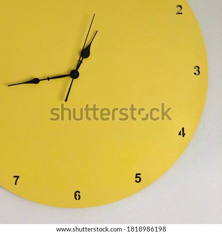 Close-up horizontal picture of a part of yellow round wall clock with hours from two to seven and  black thin vintage watch hands on the white wall