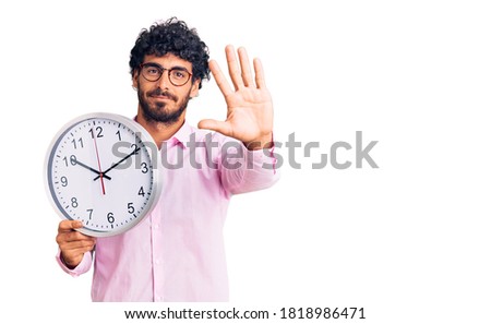 Handsome young man with curly hair and bear holding big clock with open hand doing stop sign with serious and confident expression, defense gesture 