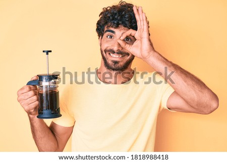 Handsome young man with curly hair and bear holding french coffee maker smiling happy doing ok sign with hand on eye looking through fingers 