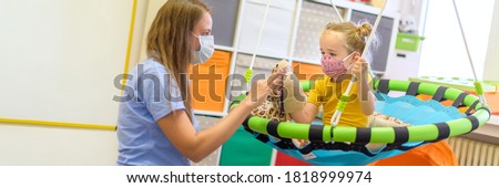 Toddler girl in child occupational therapy session doing playful exercises with her therapist during Covid - 19 pandemic, both wearing protective face masks. Web banner. Royalty-Free Stock Photo #1818999974