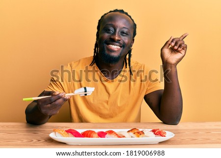 Handsome young black man eating sushi sitting on the table smiling happy pointing with hand and finger to the side 