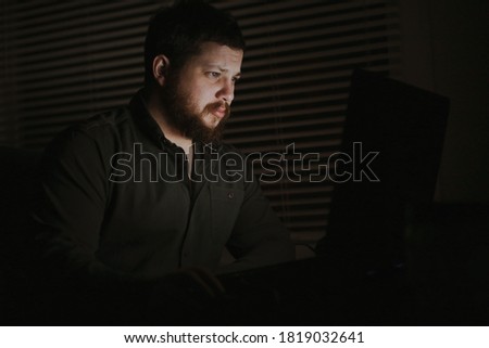 Young male caucasian programmer working with laptop lately at night, jalousie in background. Concept of smart man working at home as freelancer.