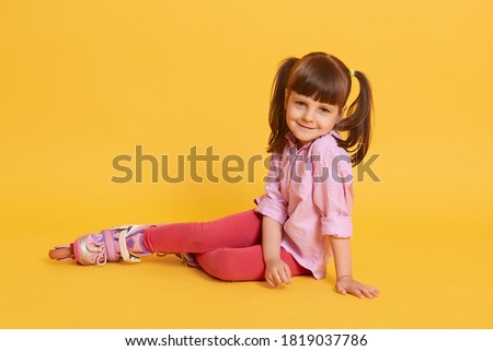 Little cute Caucasian woman with roller skates on floor, charming female kid with ponytails, wearing shirt and leggins, posing isolated over yellow background.