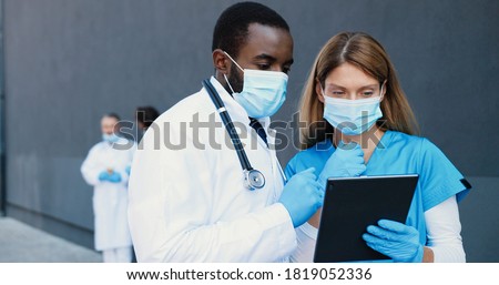 Mixed-races couple, man and woman, doctors in medical masks talking and using tablet device. Multi ethnic male and female physicians tapping and scrolling on gadget computer. Coworking in hospital. Royalty-Free Stock Photo #1819052336