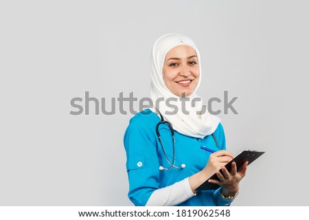 Portrait of a friendly, Muslim doctor or nurse woman in a hijab with a stethoscope writes a prescription on pills on a gray background.