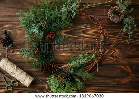 Rustic christmas wreath on wooden table flat lay, holiday advent. Modern christmas wreath with red berries, cedar branches and pine cone on rustic table with scissors and twine. Festive workshop
