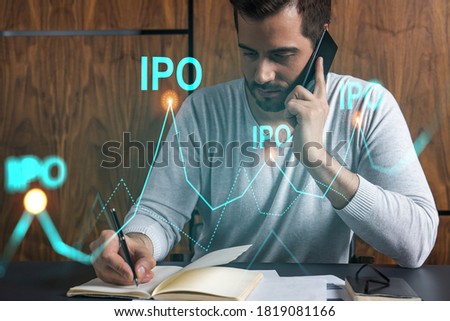 Handsome analyst in casual long sleeve talking phone, taking notes at office workplace try to analyze IPO project. Double exposure. Initial public offering hologram. Royalty-Free Stock Photo #1819081166