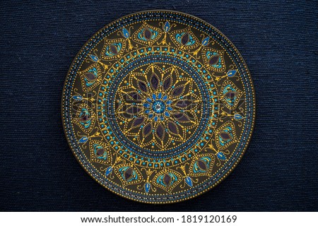 Decorative ceramic plate with black, blue and golden colors, painted plate on background of fabric, closeup, top view. Decorative porcelain plate painted with acrylic paints, handwork, dot painting