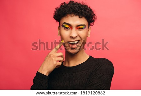 Gay man wearing multicolored shadows on the eyelids winking at camera. Happy transgender male winking an eye against red background. Royalty-Free Stock Photo #1819120862