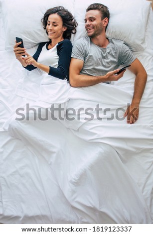 Top view photo of the beautiful woman and the handsome young man are using smartphones while lying in a big white bed. Chatting online. The photo above of the couple.