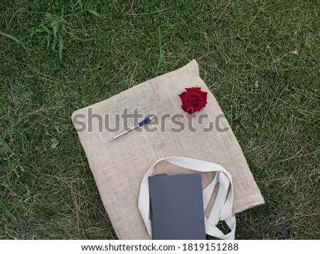 A maroon rose, a black notebook, a metal pen, and a light beige linen makeshift bag lie on the green grass on a Sunny summer day.