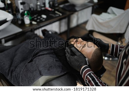 moisturizing the skin with a hot black towel after shaving, skin care. Professional work of a master in a hair salon. Royalty-Free Stock Photo #1819171328