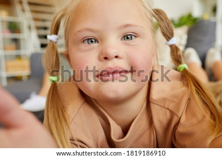 Close up portrait of blonde girl with down syndrome looking at camera while taking selfie photo at home