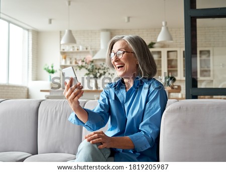 Happy mature old 60s woman holding smartphone using mobile phone app for video call, laughing while watching funny video, feeling excited winning online lottery bid on cellphone sits on couch at home. Royalty-Free Stock Photo #1819205987