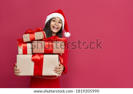 Excited funny indian latin kid child girl wears santa hat looking at camera holding many gifts boxes celebrating happy 2022 New Year isolated on red background. Merry Christmas presents shopping sale.