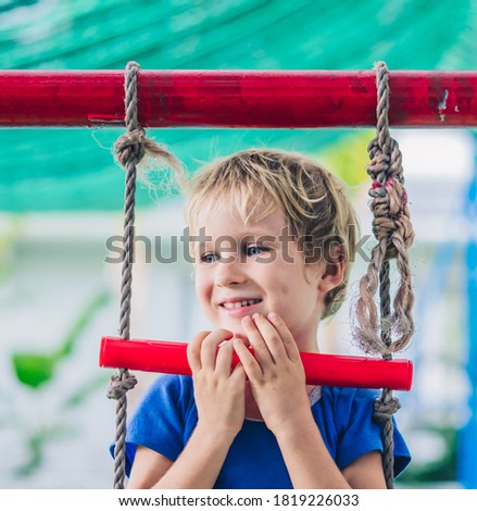 Close portrait Happy smile freckle blue eyed cute blond little boy sitting on a rope ladder, playing outside on playground in kindergarten. Daycare, activity, simple joys of childhood, summer