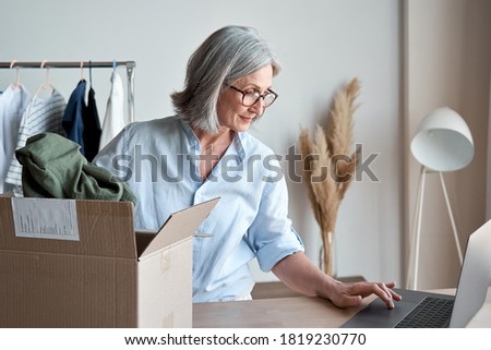 Mature woman fashion designer, dropshipping online clothing store small business owner looking for new e commerce order on laptop preparing packing in postal mail delivery shipping box at workplace. Royalty-Free Stock Photo #1819230770